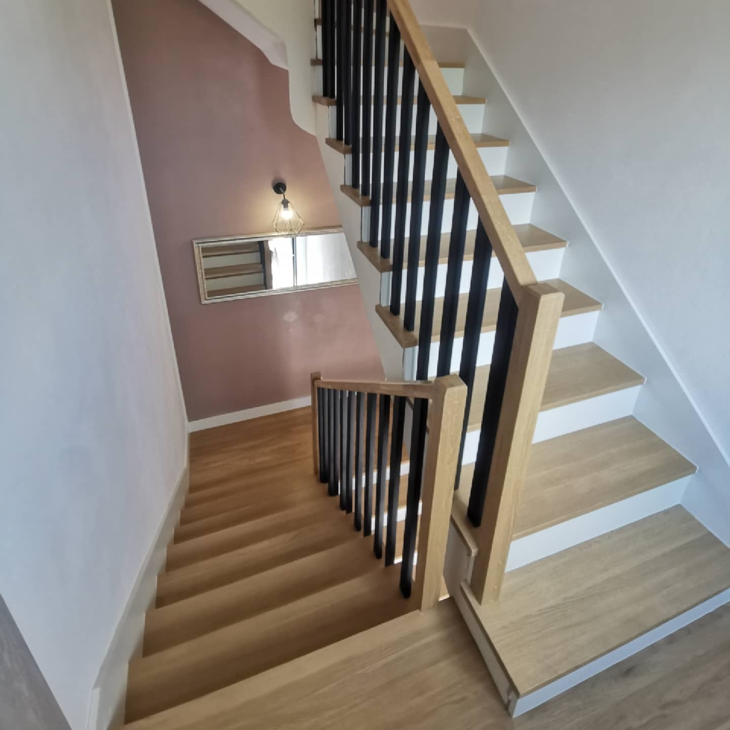 Jasne schody z drewnianymi stopniami i czarnymi balustradami, widok z góry, ściana w kolorze pudrowego różu z lustrem.