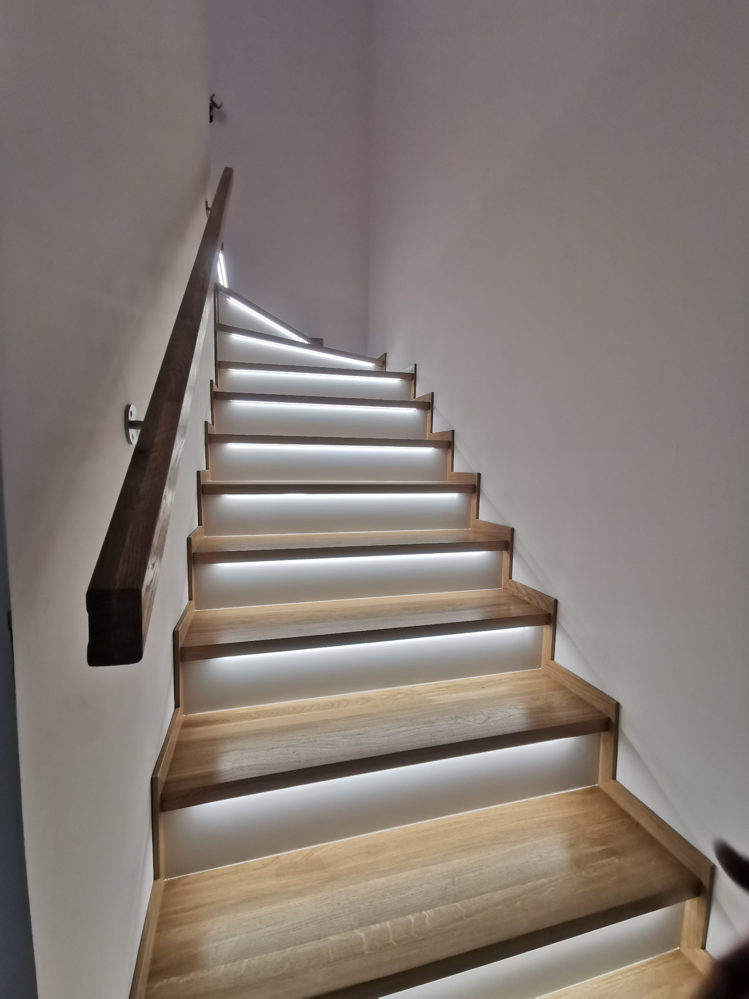 Nowoczesne schody z jasnego drewna z podświetleniem LED pod każdym stopniem, widok od dołu, minimalistyczna balustrada z ciemnego drewna.