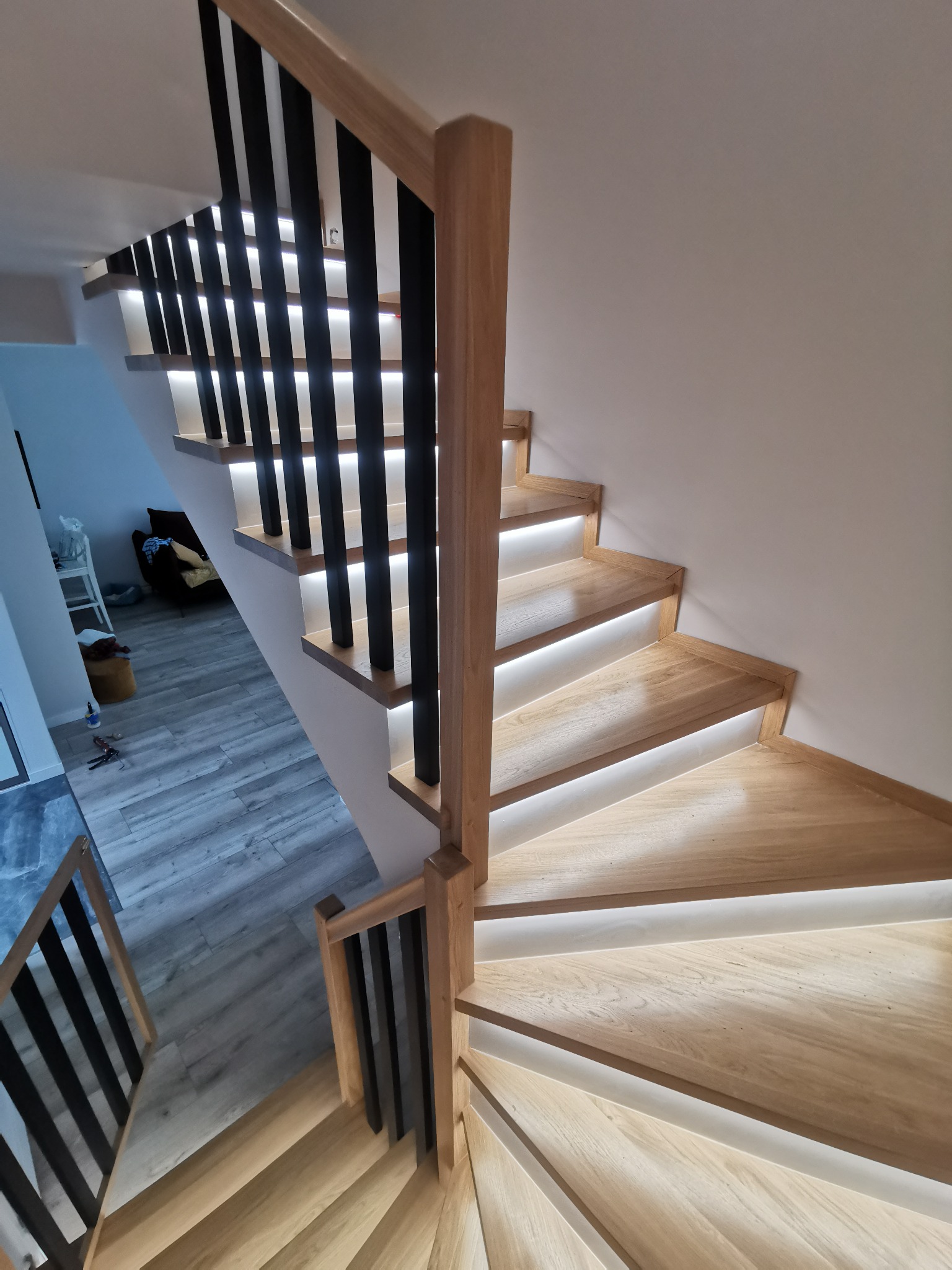 Nowoczesne schody z drewna w Sieniawie z podświetleniem LED pod każdym stopniem i czarną balustradą, widok z góry na zakręcające stopnie.