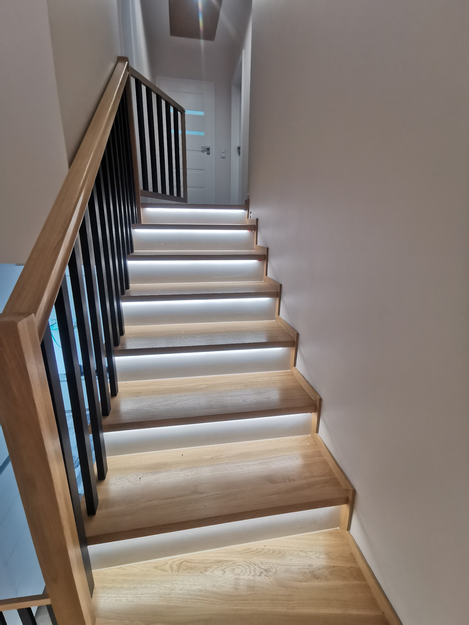 Nowoczesne schody z podświetleniem LED pod każdym stopniem, drewniane stopnie i czarna balustrada z drewnianym poręczą, widok od dołu.