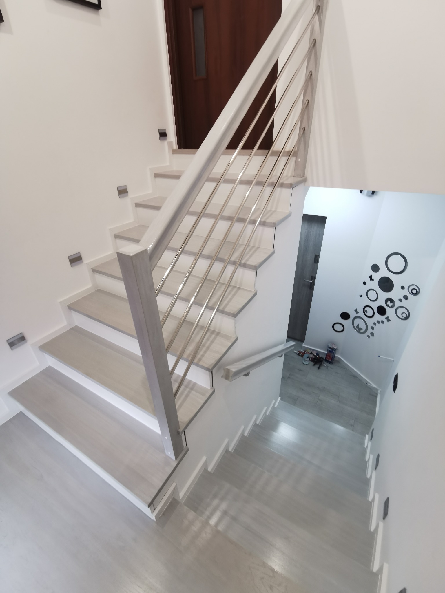 Bieg schodów z jasnego drewna z metalową balustradą, widok z góry, na ścianie dekoracje w postaci czarnych kółek.