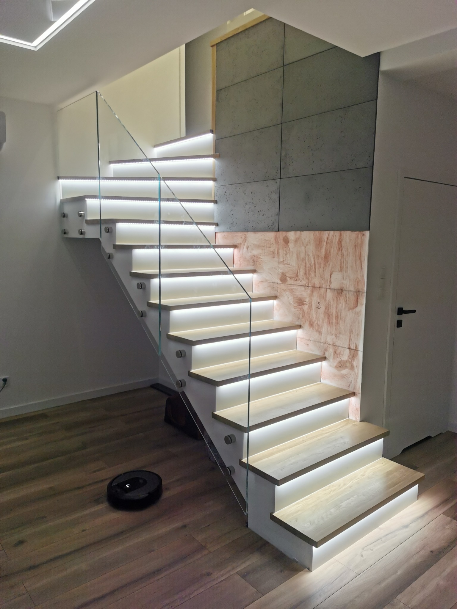 Nowoczesne schody drewniane z podświetleniem LED i szklaną balustradą, robot sprzątający na drewnianej podłodze.