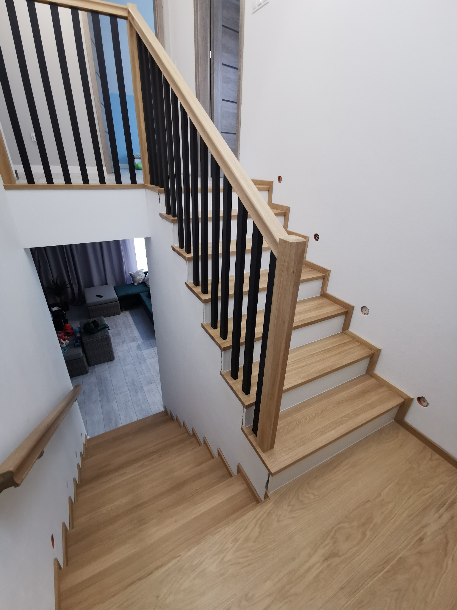 Jasne, dębowe stopnie schodów z czarną, pionową balustradą, widok z góry na dół, z widocznym fragmentem salonu w tle.