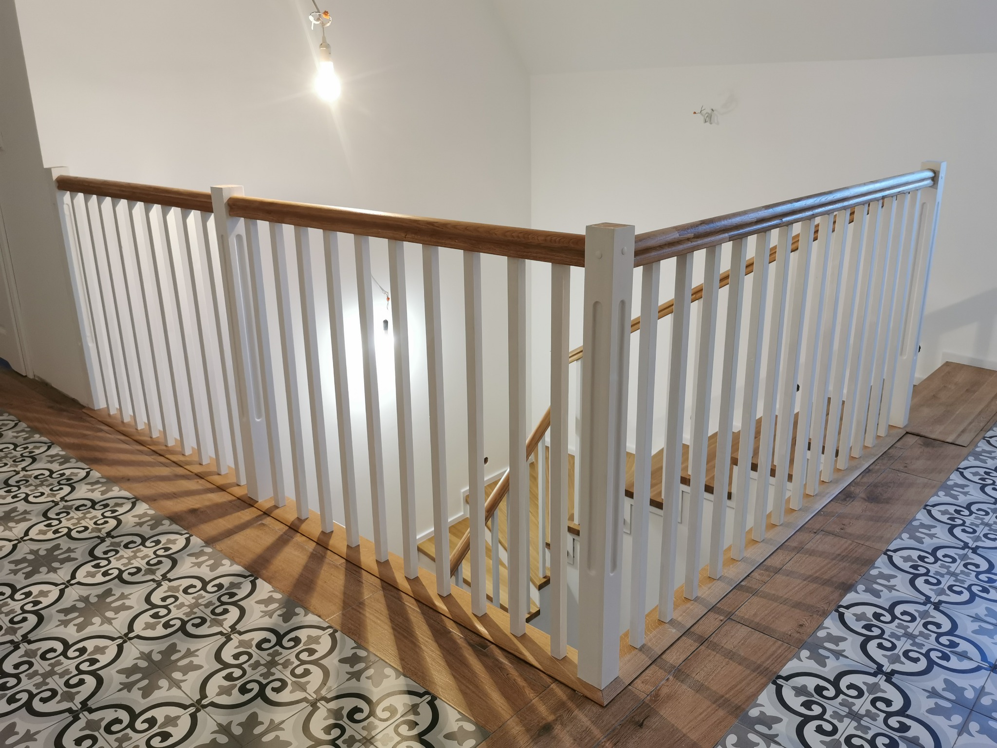 Fragment klatki schodowej z białą balustradą z drewnianym poręczą, widoczne schody i wzorzysta podłoga.