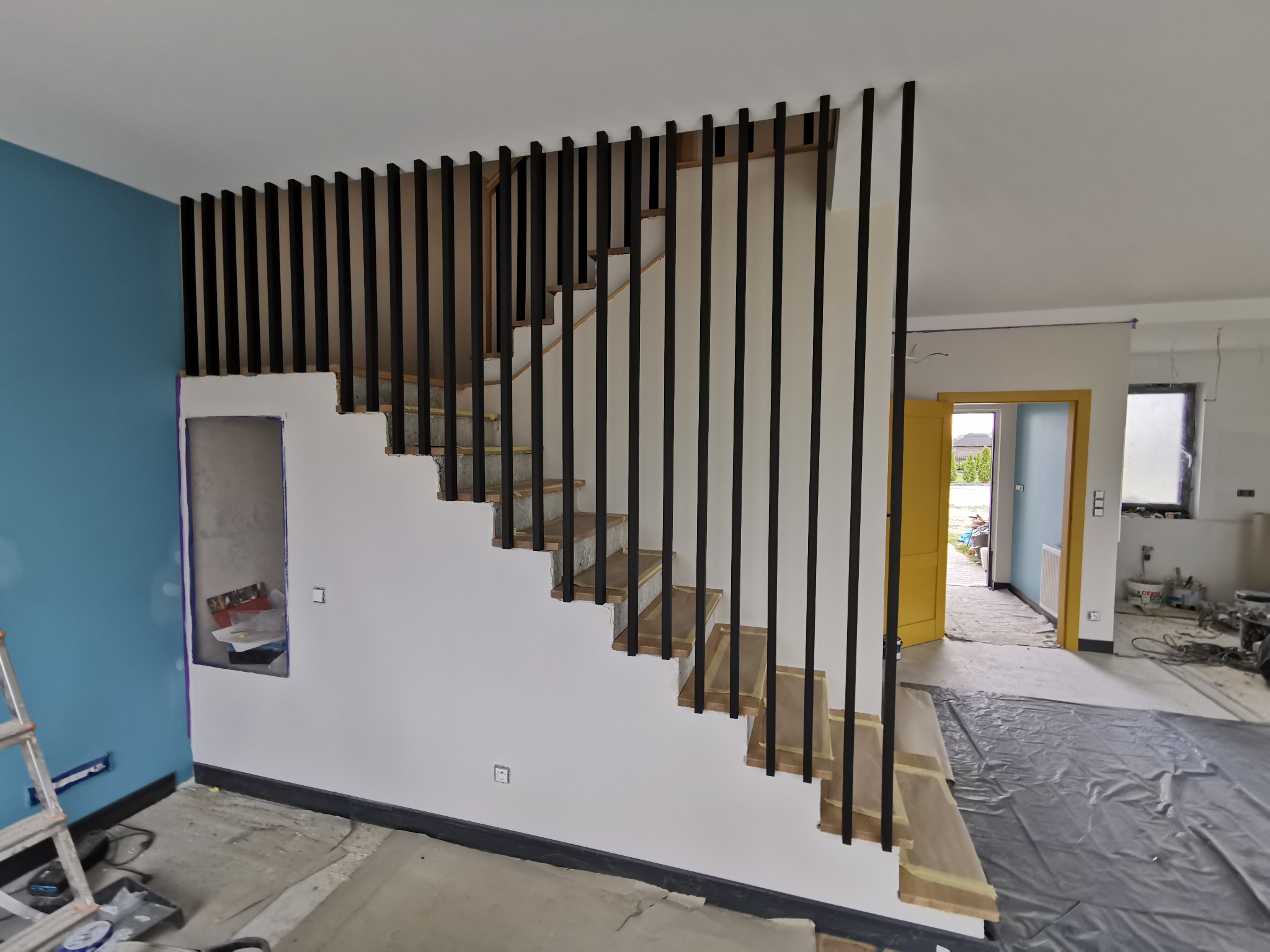 Nowoczesne schody z drewnianymi stopniami i czarną, pionową balustradą w trakcie montażu, widoczne zabezpieczenia na stopniach, w tle pomieszczenie w stanie surowym.
