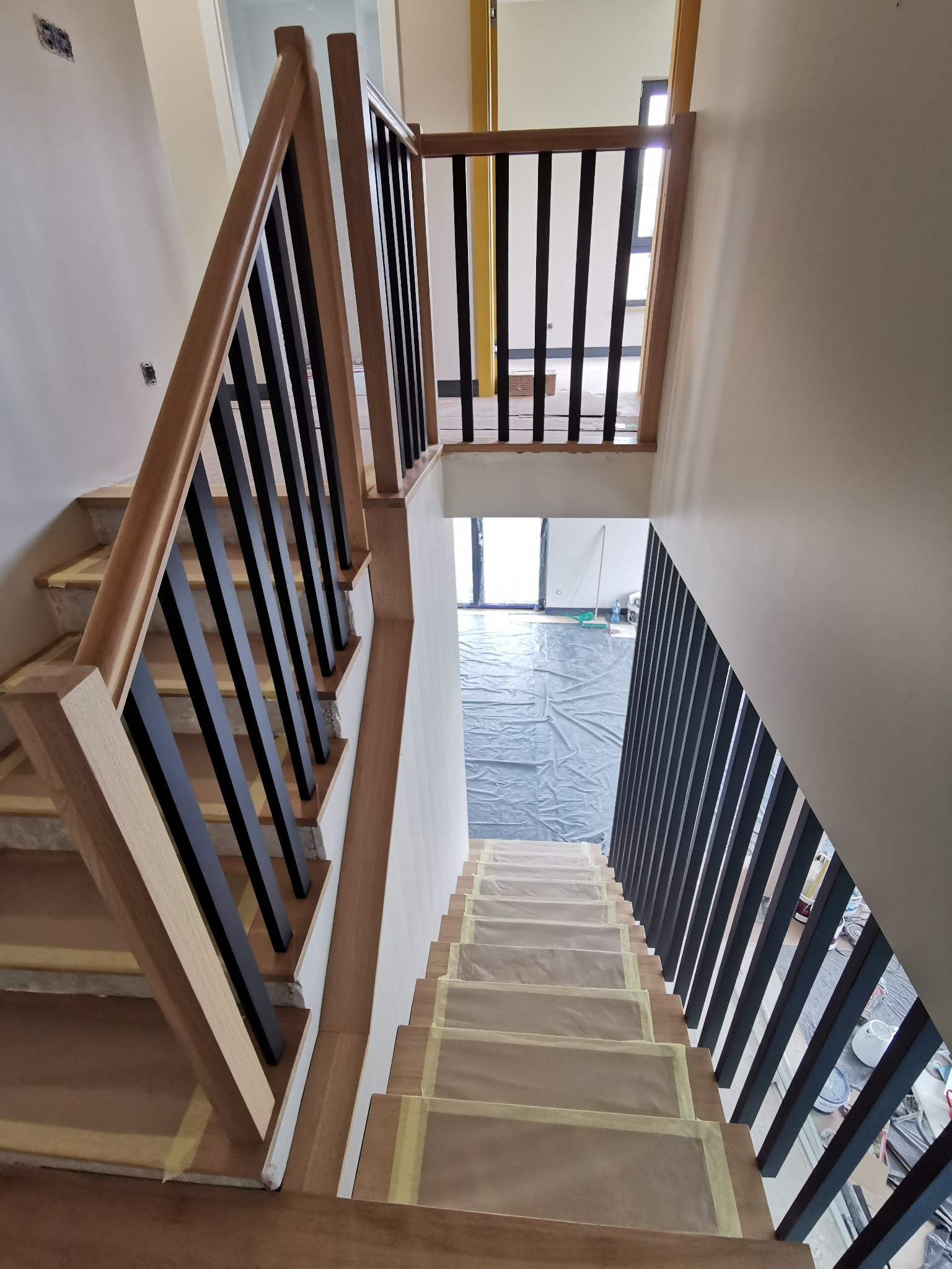 Widok z góry na drewniane schody z czarnymi, pionowymi balustradami, zabezpieczone folią i taśmą malarską podczas remontu.