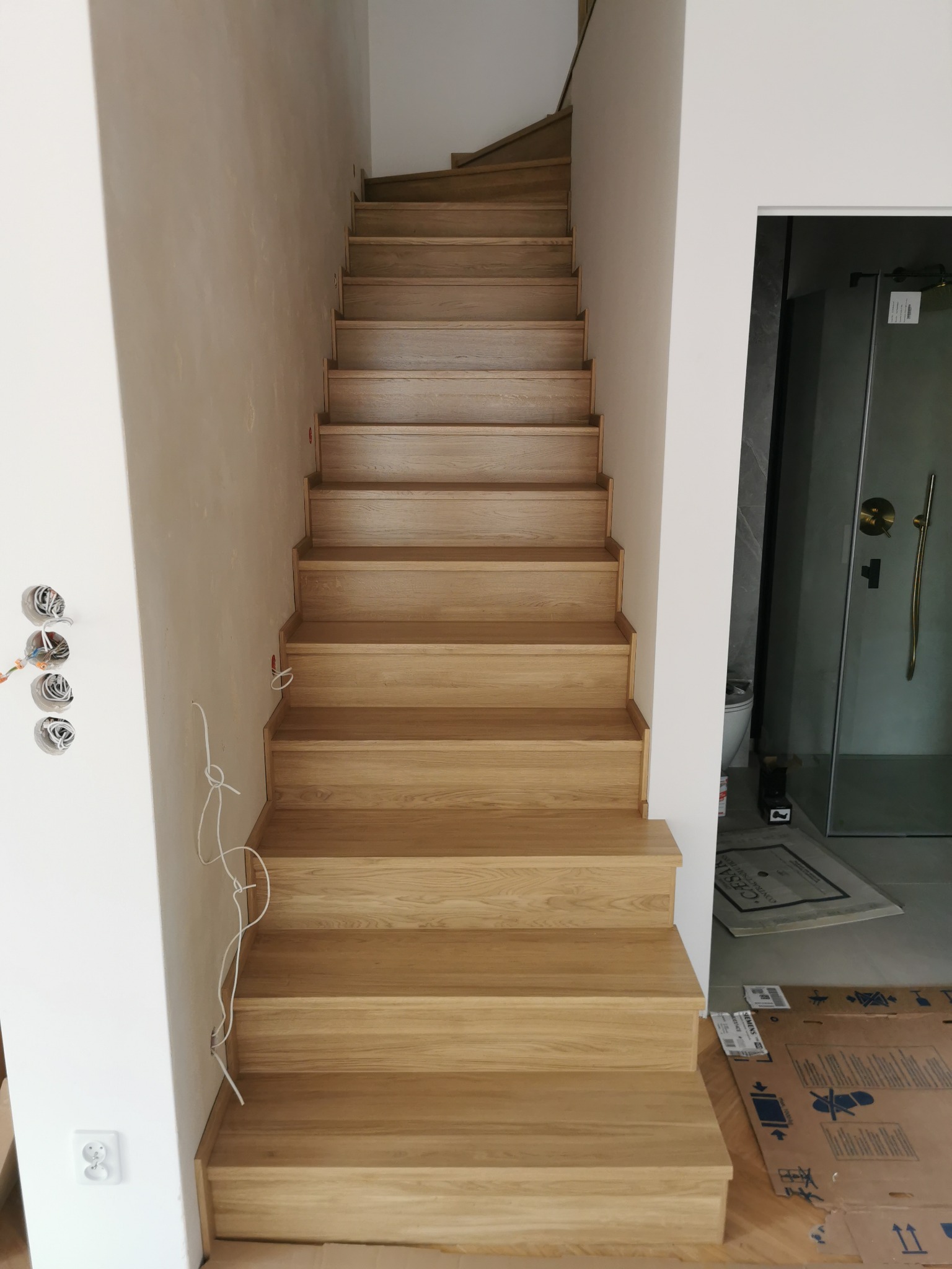 Jasne, dębowe stopnie schodów w nowym domu, widoczne przygotowania do instalacji elektrycznej i wejście do łazienki z kabiną prysznicową.