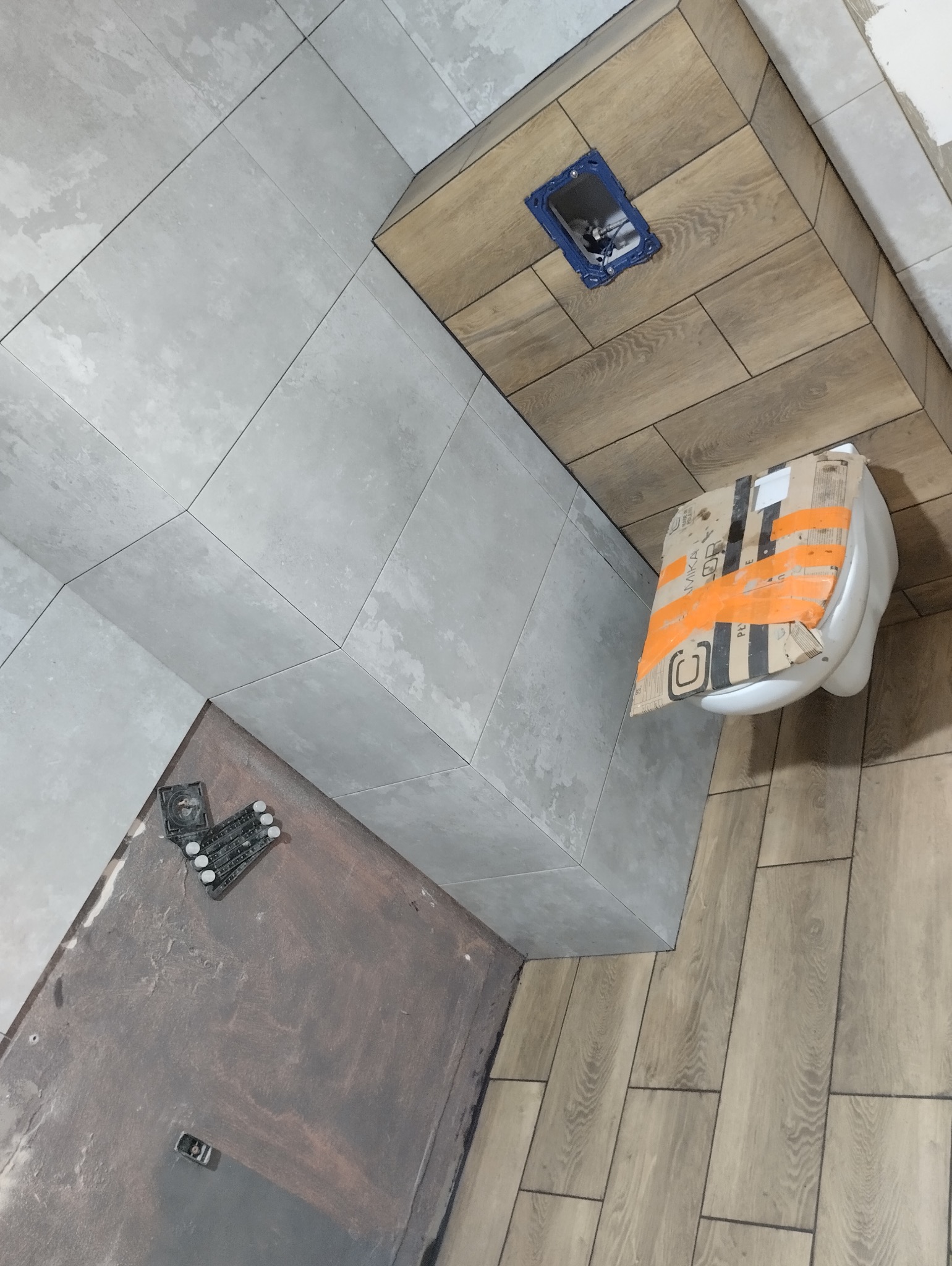 Łazienka w trakcie remontu: szare płytki na ścianach ułożone w geometryczny wzór, drewnopodobne płytki na podłodze, częściowo zamontowana toaleta w kartonie, widoczne narzędzia glazurnicze.