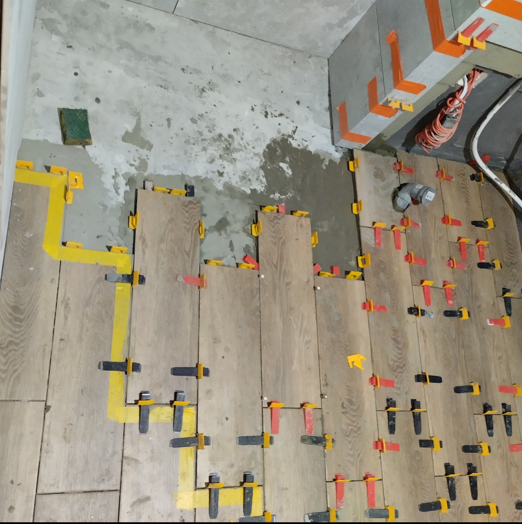 Układanie płytek imitujących drewno na podłodze, widoczne systemy poziomowania płytek w kolorach żółtym, czerwonym i czarnym, fragment surowej ściany i podłogi z widocznym odpływem.