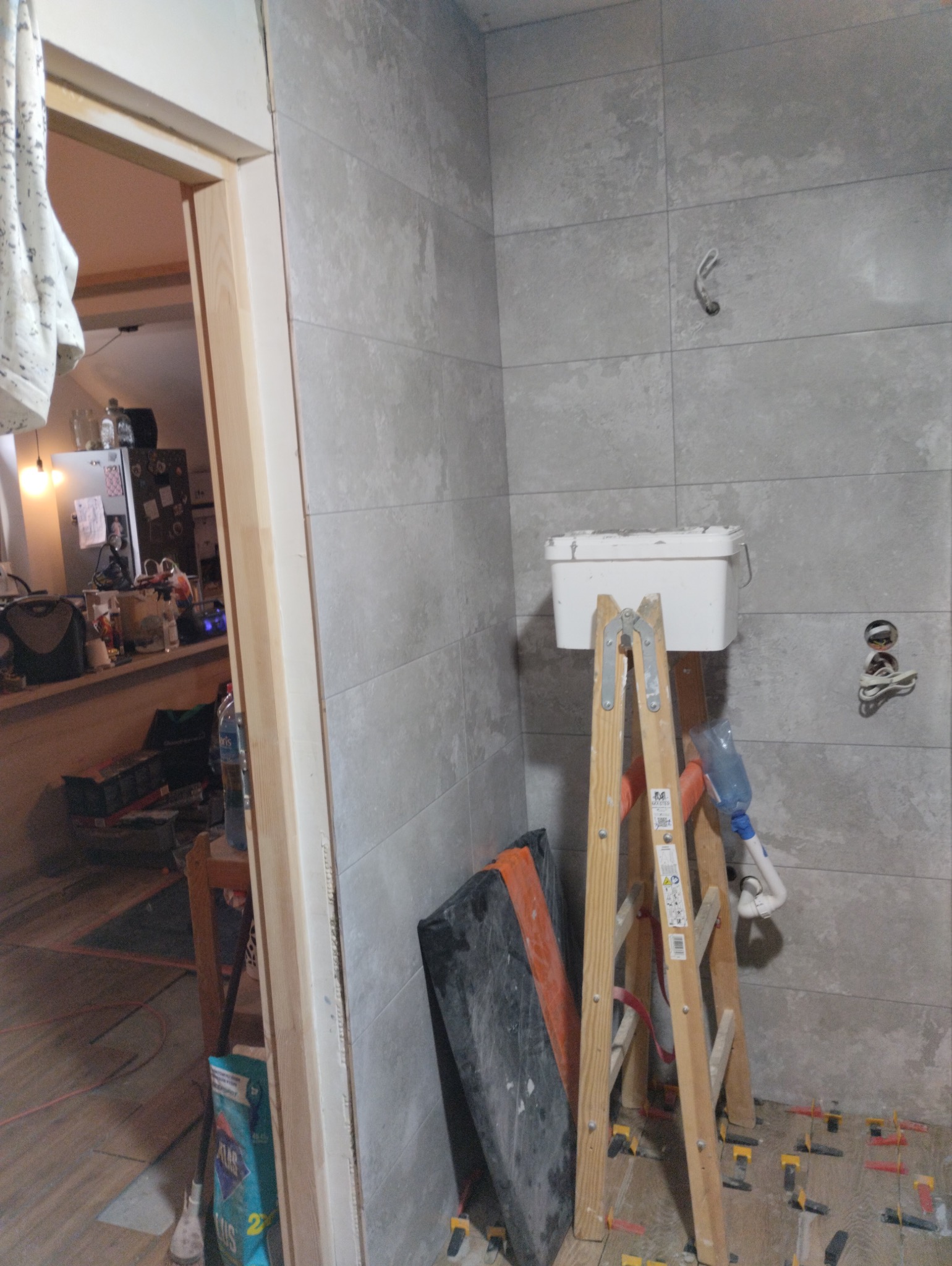 Szare płytki ceramiczne na ścianie łazienki, drabina malarska z wiadrem, widoczne krzyżyki dystansowe na podłodze, fragment ościeżnicy drzwiowej i widok na kuchnię w tle.