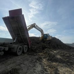 Mariusz Ślęzak Sklep Spożywczo-Przemysłowy - Żółta koparka na hałdzie ziemi i wywrotka z podniesioną skrzynią w trakcie prac ziemnych na tle błękitnego nieba z chmurami.