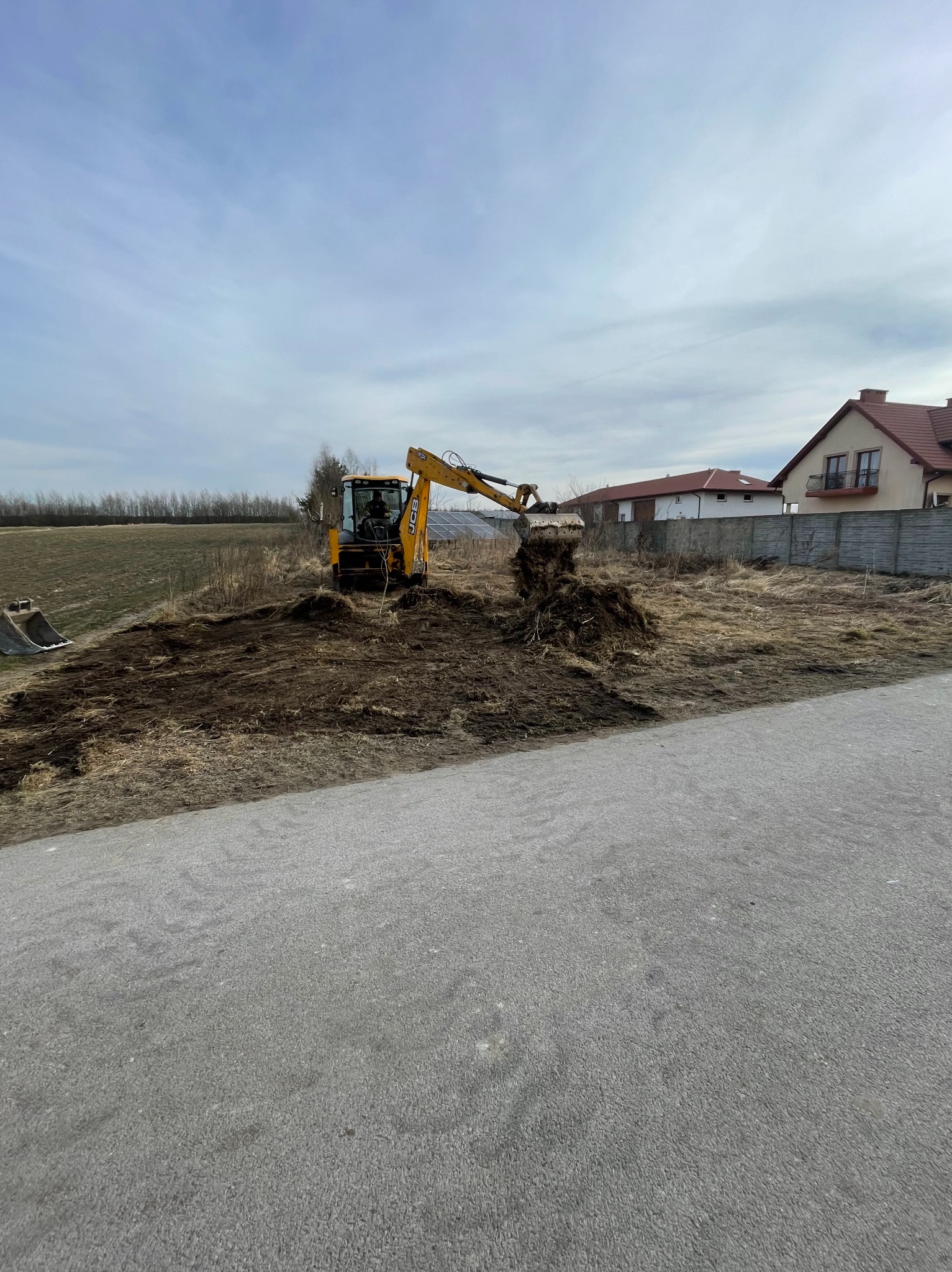 Żółta minikoparka JCB podczas prac ziemnych na działce, z widocznym wykopem i stertą ziemi, w tle domy jednorodzinne i pochmurne niebo.