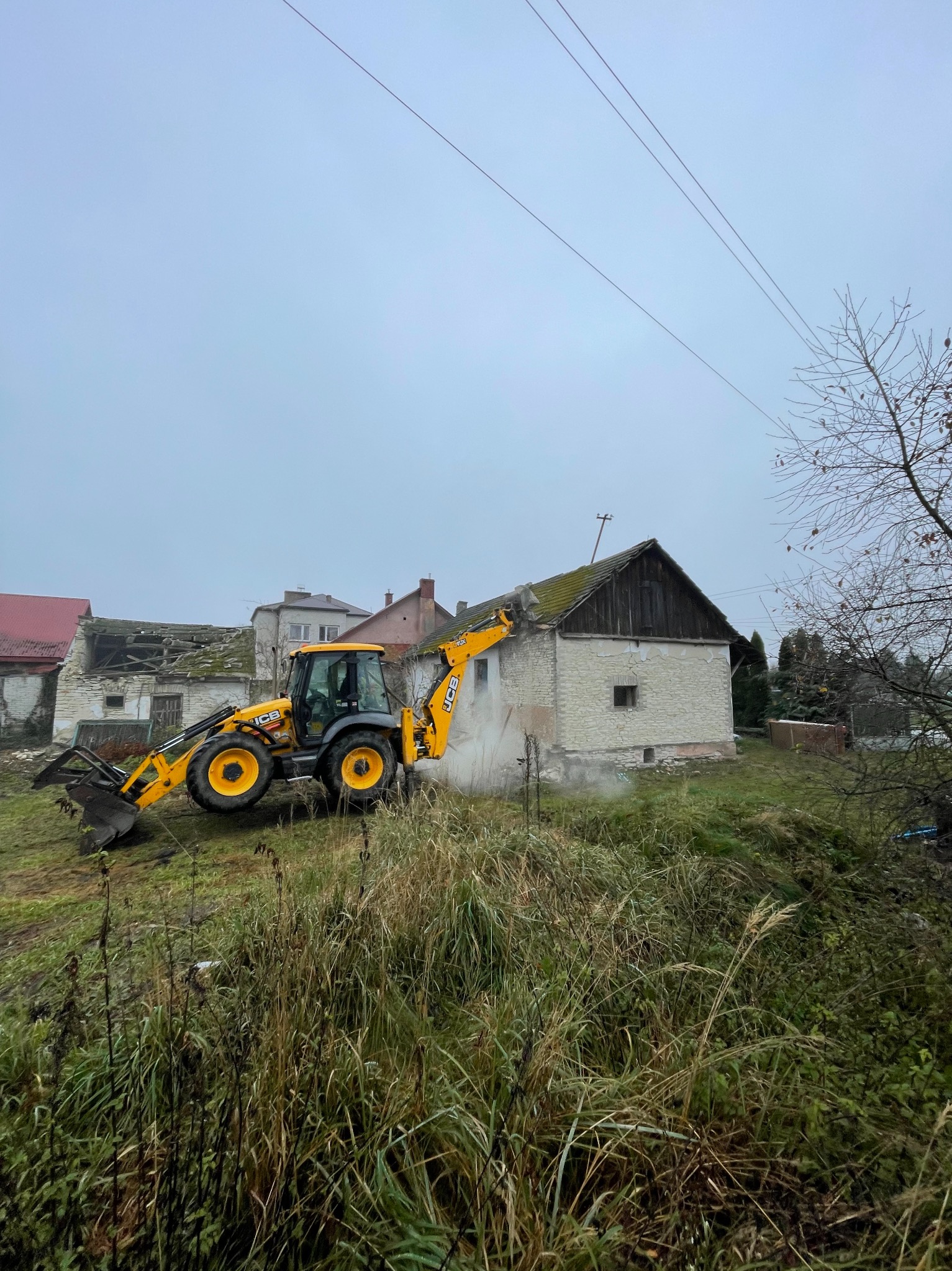 Żółta koparko-ładowarka JCB podczas częściowego demontażu murowanego budynku gospodarczego z drewnianą nadbudówką, widoczne tumany kurzu, na drugim planie zabudowania wiejskie i słupy energetyczne.