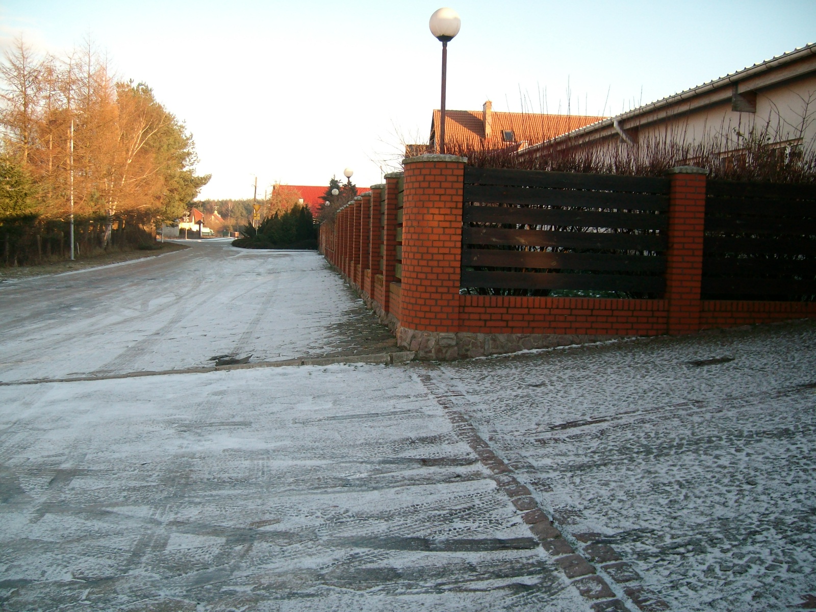 Zimowy krajobraz z ceglanym ogrodzeniem z drewnianymi panelami i lampami ulicznymi, częściowo pokryty śniegiem.
