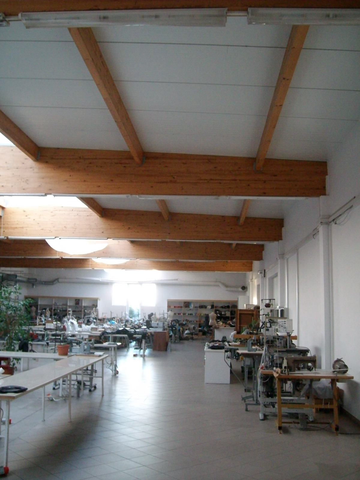 Przestronna hala produkcyjna szwalni z drewnianą konstrukcją stropu, maszynami do szycia, stołami roboczymi i regałami z akcesoriami.