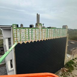 Clover - Budynek w trakcie ocieplania: widoczna konstrukcja drewniana z izolacją, częściowo pokryta czarnym materiałem elewacyjnym, z fragmentem rusztowania i widokiem na okolicę.