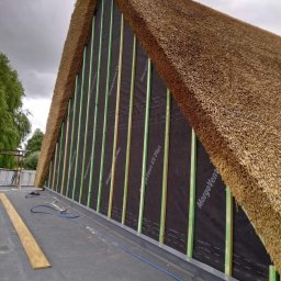 Clover - Fragment dachu krytego strzechą w trakcie montażu, widoczne deski konstrukcyjne, membrana i elementy mocujące, pochmurne niebo w tle.