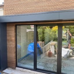 Clover - Nowoczesna zabudowa tarasu z przesuwnymi drzwiami szklanymi i poziomą okładziną drewnianą, widoczne elementy instalacji grzewczej w podłodze i odbicie osoby pracującej na zewnątrz.