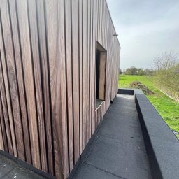 Clover - Elewacja budynku wykończona pionowymi panelami z ciemnego drewna, widok z poziomu dachu pokrytego czarną membraną, w tle zielone pole i pochmurne niebo.