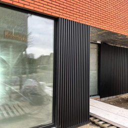 Clover - Elewacja budynku z cegły klinkierowej z czarnymi, pionowymi panelami tworzącymi nowoczesny akcent architektoniczny, widoczne okno z odbiciem otoczenia, teren budowy z płytami betonowymi.