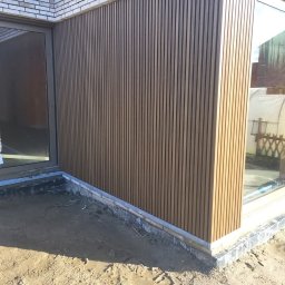 Clover - Montaż pionowych paneli elewacyjnych w kolorze jasnego drewna na ścianie domu, widoczne okna z szarymi ramami i przygotowany teren wokół budynku.