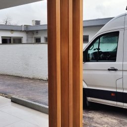 Clover - Nowoczesny taras z drewnianymi słupami pod zadaszeniem, obok biały bus, widoczne czarne rury w ziemi, w tle biały budynek.