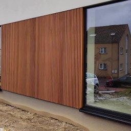 Clover - Nowoczesna elewacja domu z pionowymi panelami w kolorze drewna, widoczne duże okna z czarnymi ramami i fragment terenu przed budynkiem w trakcie prac wykończeniowych.