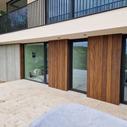 Clover - Nowoczesny taras z płyt kamiennych, zabudowany przesuwnymi panelami z drewna egzotycznego, widok na zieleń za oknem, częściowo przykryty mebel ogrodowy w rogu.