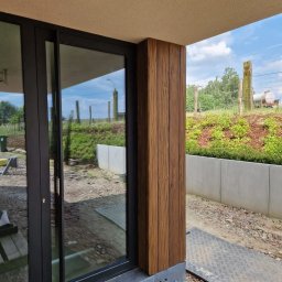 Clover - Nowoczesny dom w Kapałowie z przesuwnymi drzwiami tarasowymi i obudową słupa z paneli drewnopodobnych, widok na ogród.