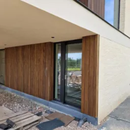 Nowoczesny budynek z elewacją z jasnej cegły i okładziną z paneli drewnopodobnych, widoczne narzędzia i materiały budowlane sugerujące trwające prace wykończeniowe.