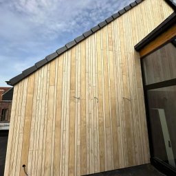 Clover - Nowoczesna elewacja domu wykończona jasnym drewnem, widoczne fragmenty dachu z ciemną dachówką i okna z czarnymi ramami, przewody na elewacji czekające na podłączenie.