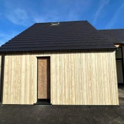 Clover - Nowo wybudowany budynek z jasną, pionową okładziną drewnianą i czarnym dachem z dachówką, widoczne okno dachowe i zabezpieczone wejście.