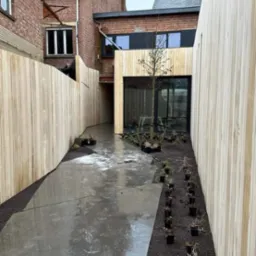 Nowoczesny ogr&oacute;d z drewnianym ogrodzeniem i elementami zabudowy, świeżo po deszczu, z rzędami małych roślin w doniczkach wzdłuż ścieżki.