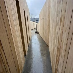 Clover - Wąskie przejście między dwiema drewnianymi ścianami z pionowych desek, mokry betonowy chodnik, pochmurne niebo w tle.