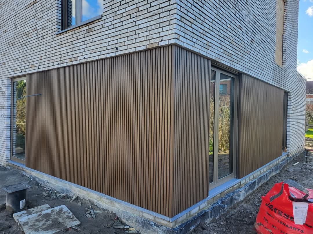 Elewacja budynku wykończona brązowymi panelami pionowymi, widoczne okna i fragment ceglanej ściany, teren wokół w trakcie prac wykończeniowych.