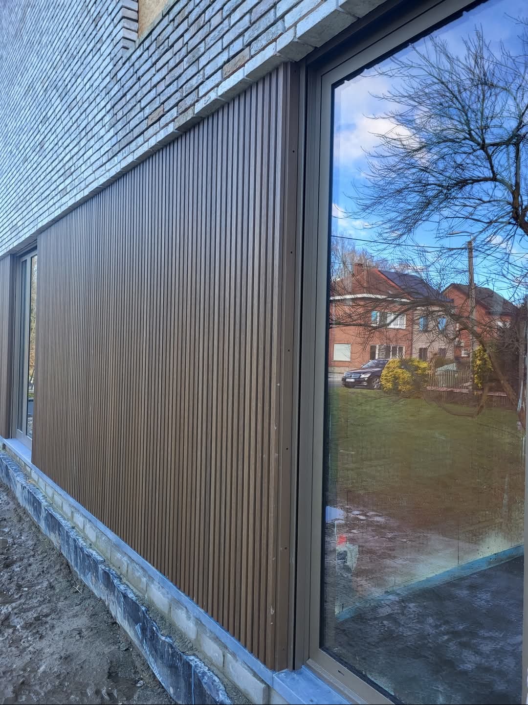 Elewacja budynku z pionowymi panelami w odcieniach brązu, widoczna ceglana ściana powyżej i duże okno z odbiciem otoczenia, w tle dom z czerwonym dachem i zielony trawnik.