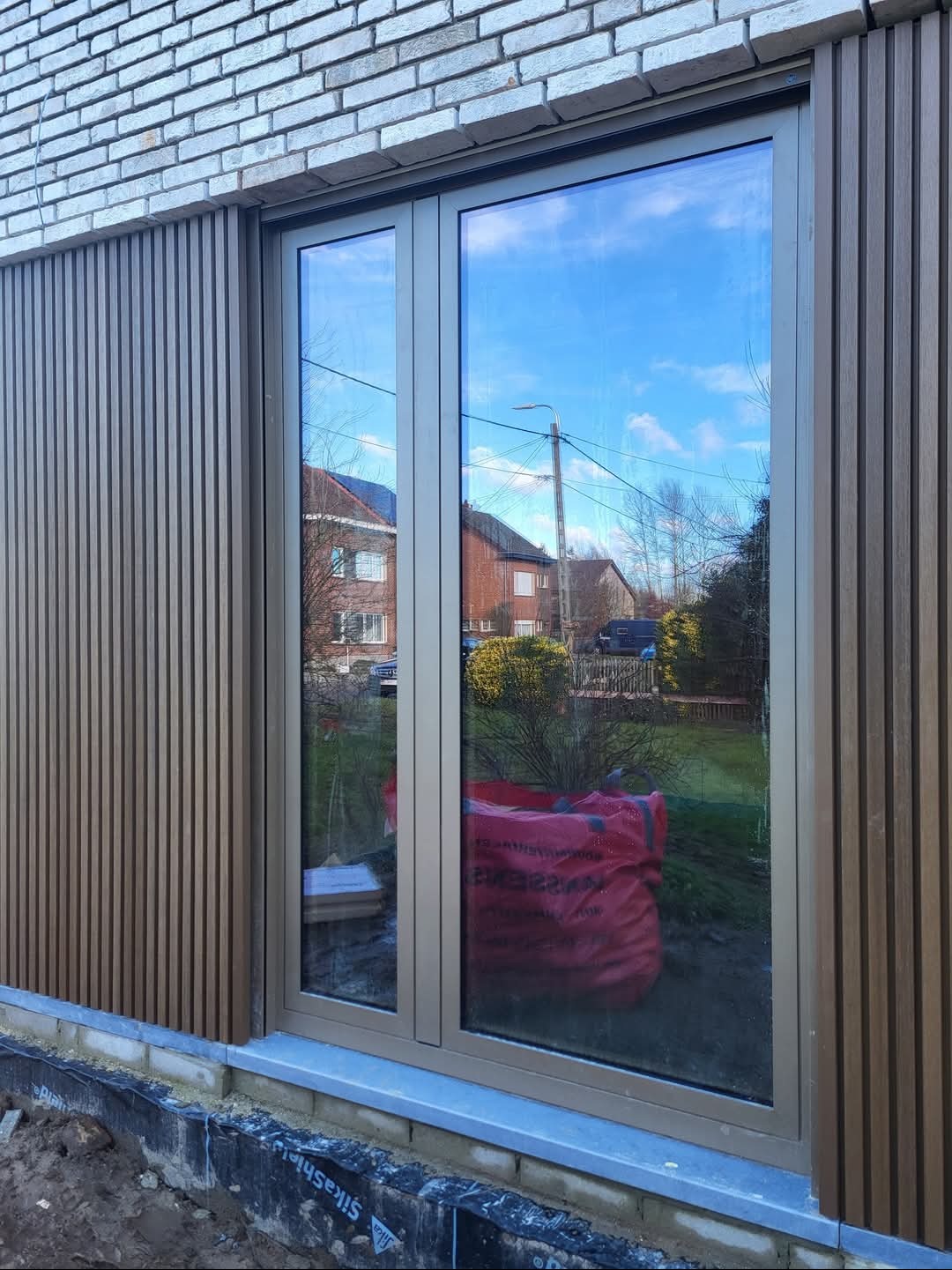 Nowoczesna elewacja z pionowymi panelami, okno z widokiem na ogród, czerwony worek na zewnątrz.