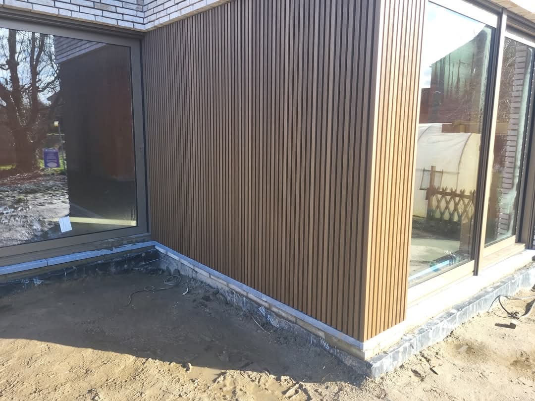 Montaż pionowych paneli elewacyjnych w kolorze jasnego drewna na ścianie domu, widoczne okna z szarymi ramami i przygotowany teren wokół budynku.