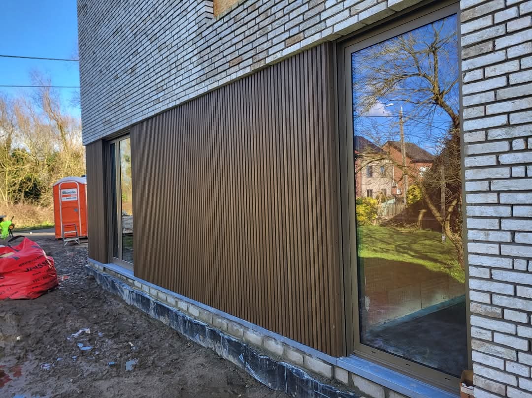 Fragment elewacji budynku z brązową okładziną z pionowych paneli, widoczne okno z odbiciem drzew i budynku, mur z jasnej cegły oraz teren budowy z workiem i przenośną toaletą.