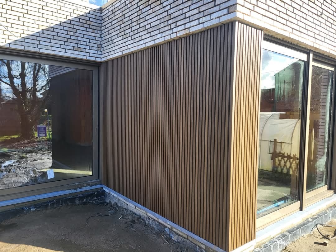 Fragment elewacji budynku z jasnej cegły i pionowo ułożonych paneli w kolorze drewna, widoczny fragment tarasu z szarym podłożem i okno z odbiciem drzewa.