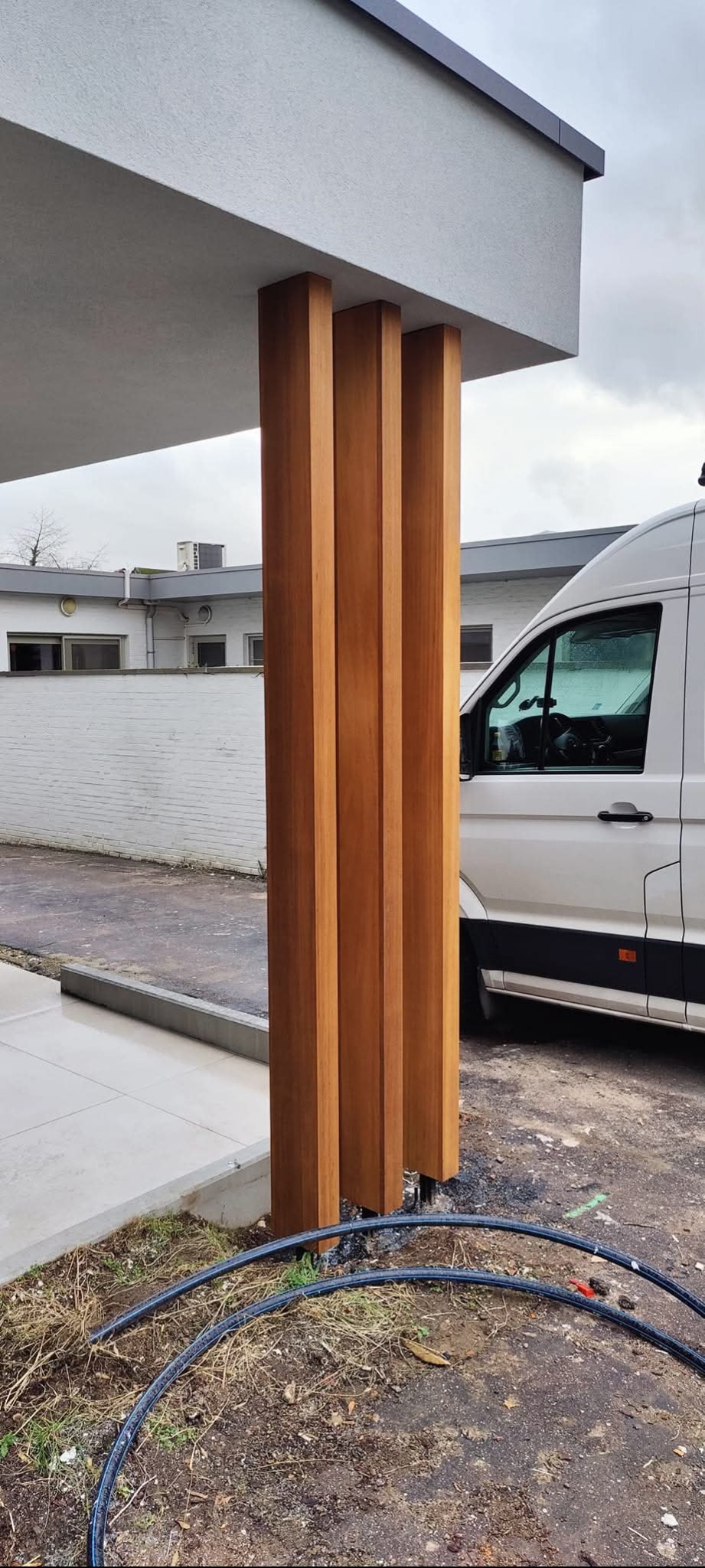 Nowoczesny taras z drewnianymi słupami pod zadaszeniem, obok biały bus, widoczne czarne rury w ziemi, w tle biały budynek.