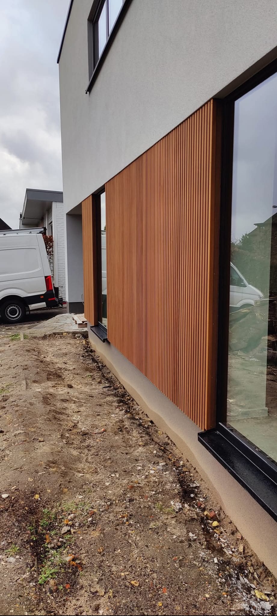 Nowoczesna elewacja budynku z drewnianą okładziną w pionowe listwy, widok z zewnątrz, z ziemią w stanie surowym i odblaskiem białego vana w oknie.