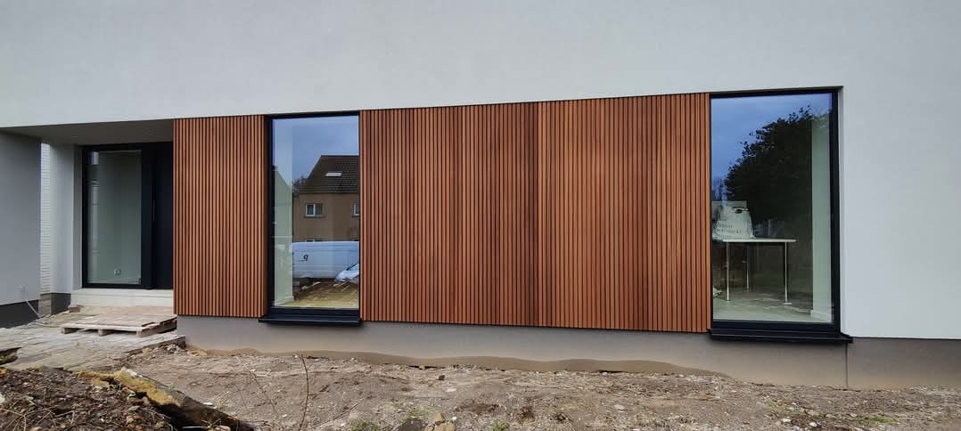 Elewacja budynku z fragmentami wykończonymi brązowymi panelami pionowymi, widoczne okna z czarnymi ramami i drzwi wejściowe, teren wokół budynku w trakcie prac wykończeniowych.