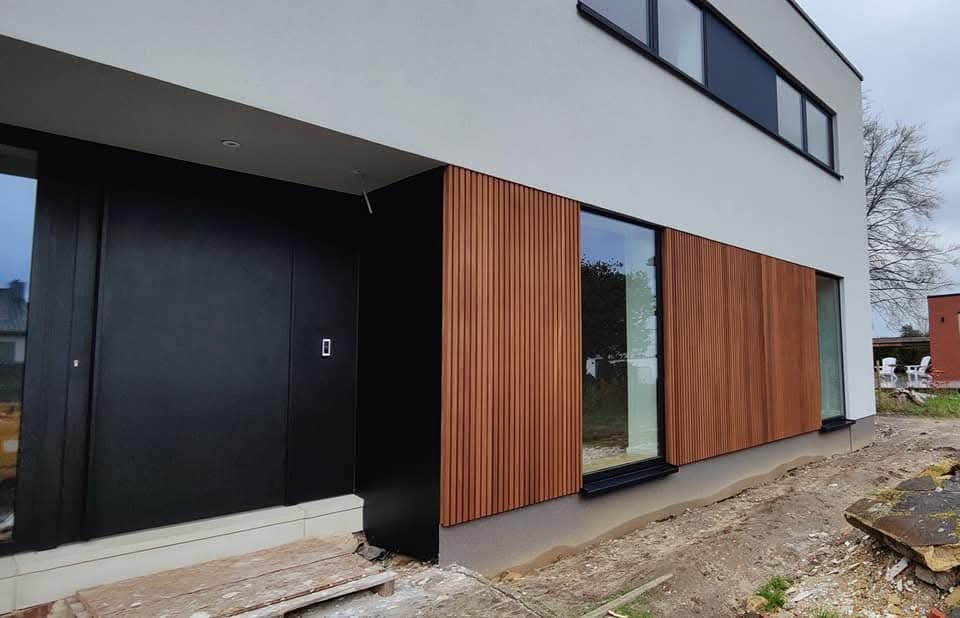Nowoczesna elewacja domu z drewnianą okładziną pionową i czarnymi elementami, widoczne okna przesuwne i niedokończone otoczenie budynku.