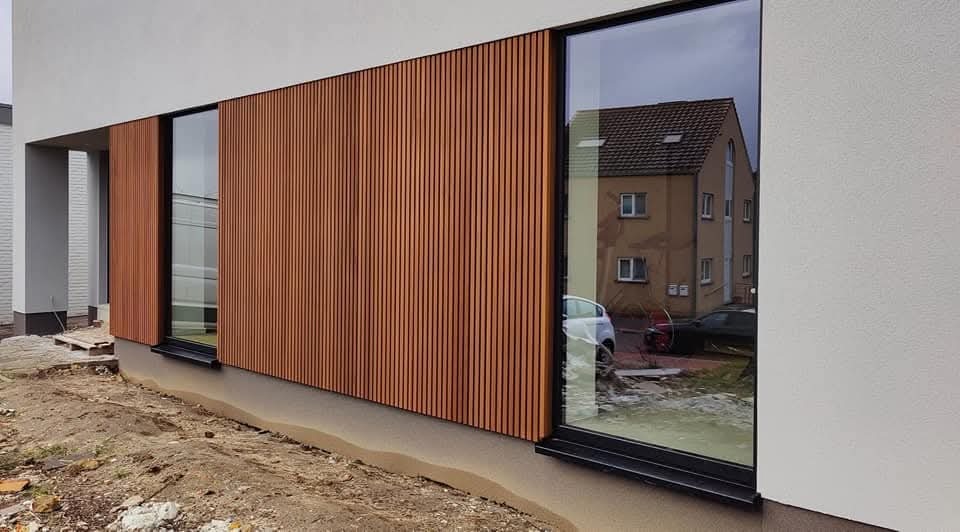 Nowoczesna elewacja domu z pionowymi panelami w kolorze drewna, widoczne duże okna z czarnymi ramami i fragment terenu przed budynkiem w trakcie prac wykończeniowych.