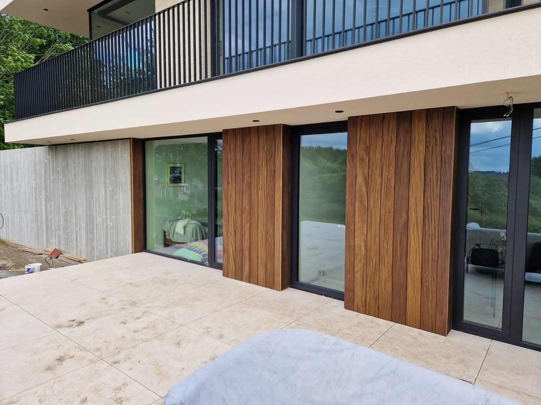 Nowoczesny taras z płyt kamiennych, zabudowany przesuwnymi panelami z drewna egzotycznego, widok na zieleń za oknem, częściowo przykryty mebel ogrodowy w rogu.