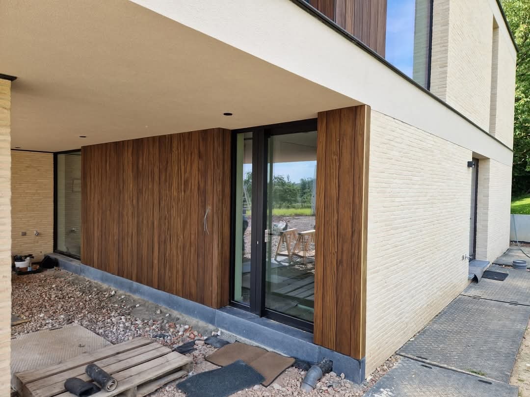 Nowoczesny budynek z elewacją z jasnej cegły i okładziną z paneli drewnopodobnych, widoczne narzędzia i materiały budowlane sugerujące trwające prace wykończeniowe.