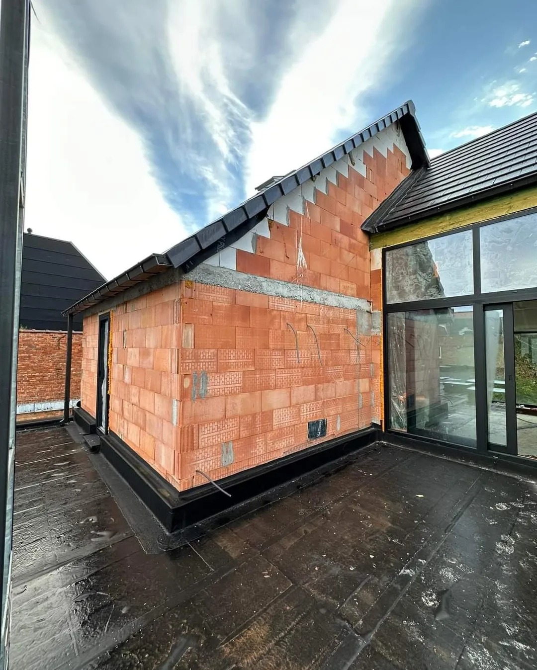 Surowy mur z czerwonej cegły porotherm, widok z czarnego dachu płaskiego, fragment okna tarasowego i niebo z chmurami w tle, budowa domu w trakcie realizacji.