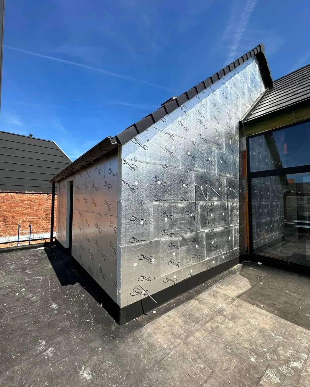 Budynek w trakcie ocieplania z zewnątrz, pokryty srebrną folią izolacyjną z nadrukowanym logo 'RECTICEL', widoczne okno i fragment dachu.