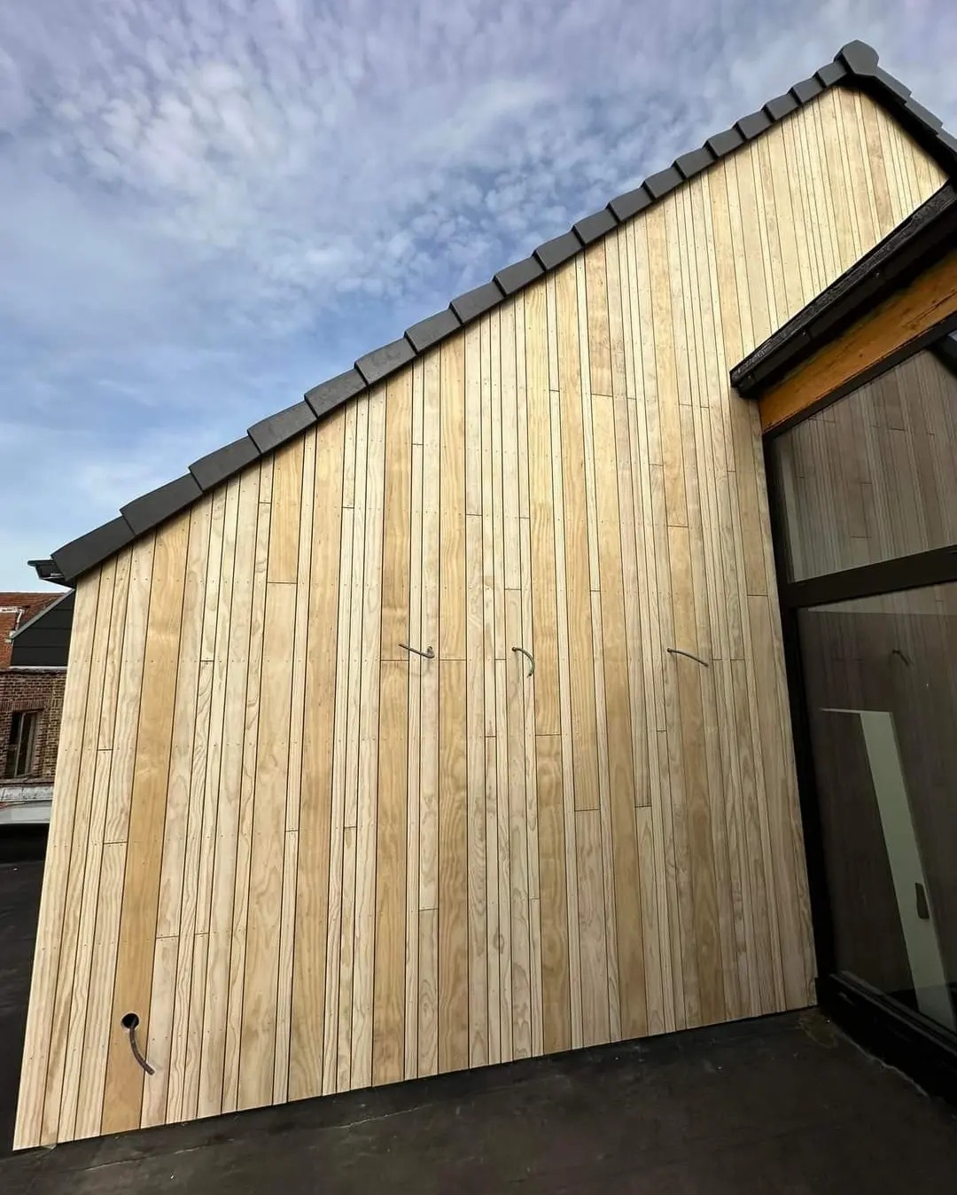 Nowoczesna elewacja domu wykończona jasnym drewnem, widoczne fragmenty dachu z ciemną dachówką i okna z czarnymi ramami, przewody na elewacji czekające na podłączenie.
